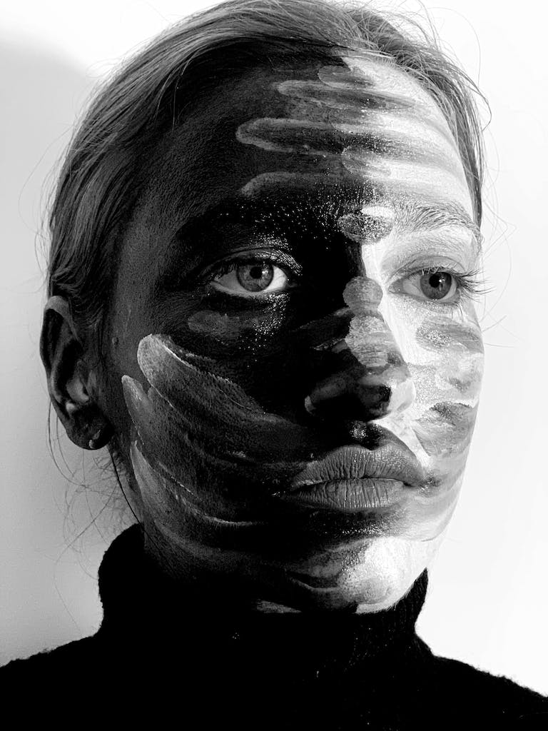 Black and white of serious young woman with dark and light dyes on face looking away pensively in studio